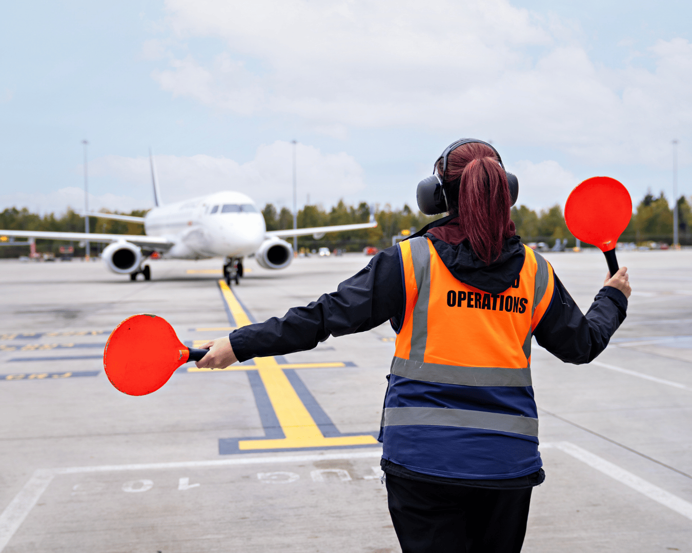 A ground crew member in an orange vest labeled "Operations" signals an incoming airplane with red paddles. The runway is clear and the sky is partly cloudy.