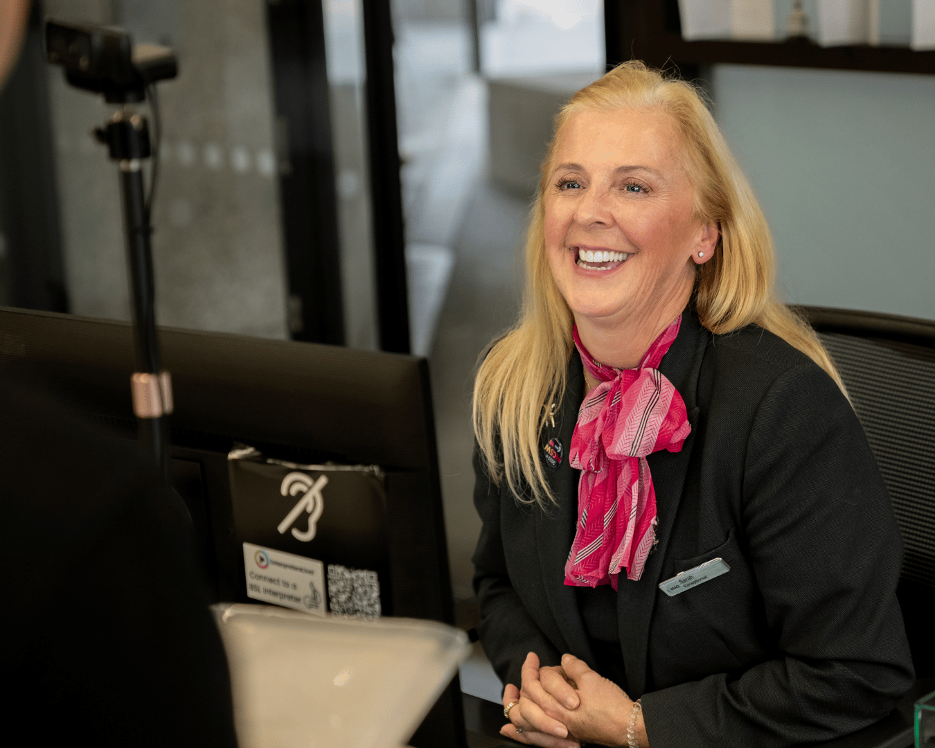 A smiling receptionist with long blonde hair and a pink scarf sits at a desk, welcoming a visitor.