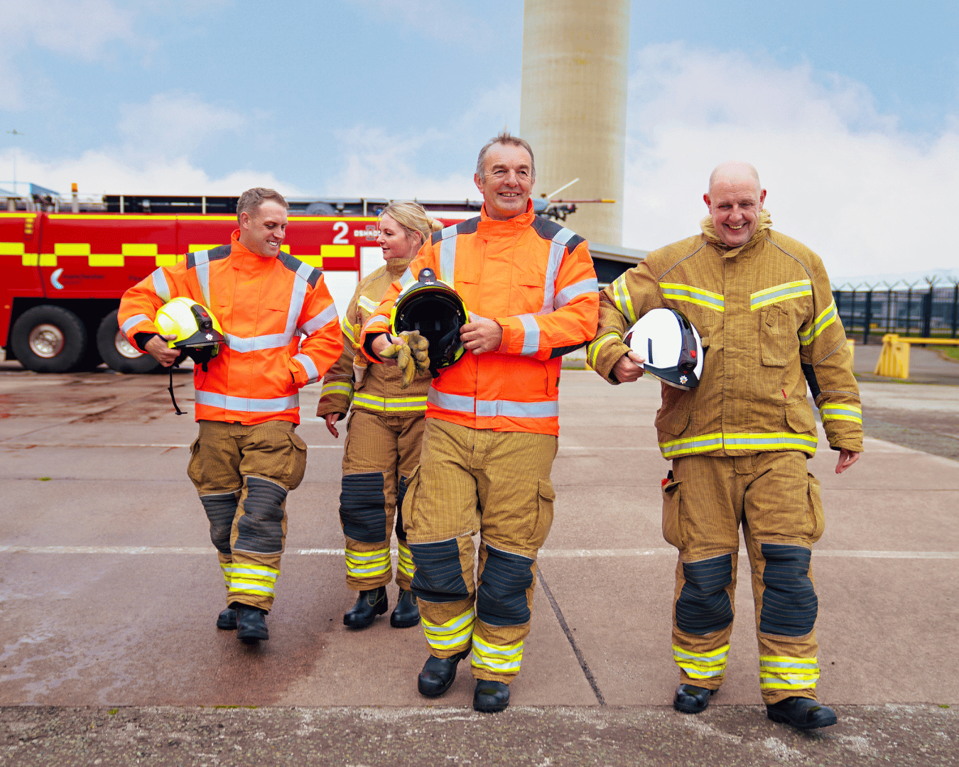 Four firefighters in bright orange jackets and protective gear walk confidently, holding helmets. A fire truck is visible in the background at an airport.