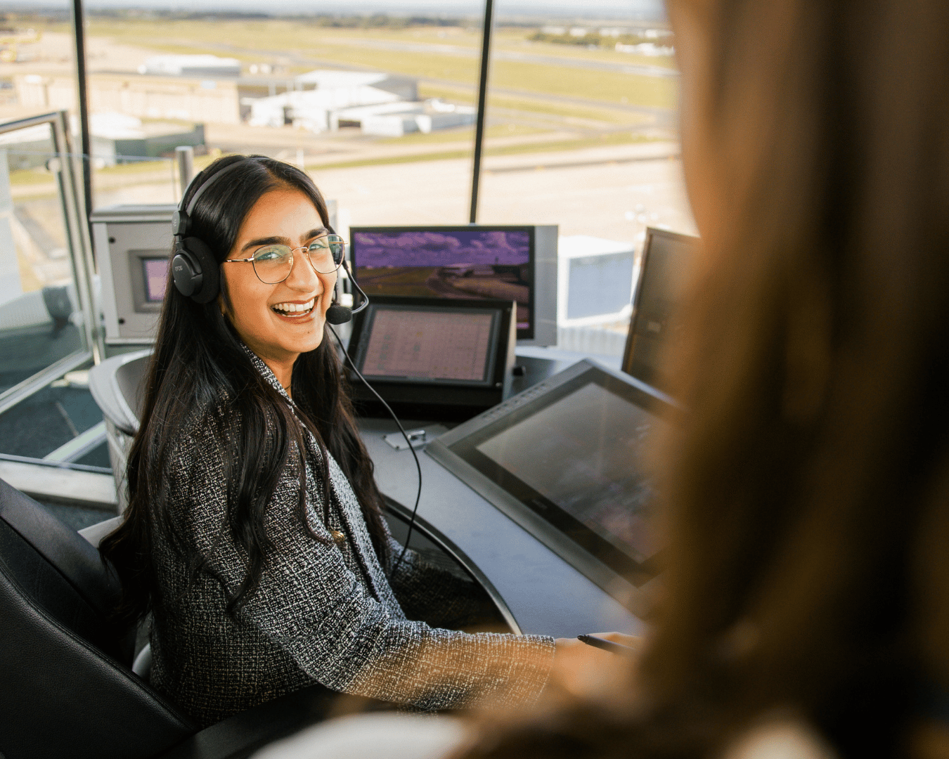 A smiling air traffic controller with long dark hair and glasses wears a headset while working at control tower monitors. Runways are visible outside.