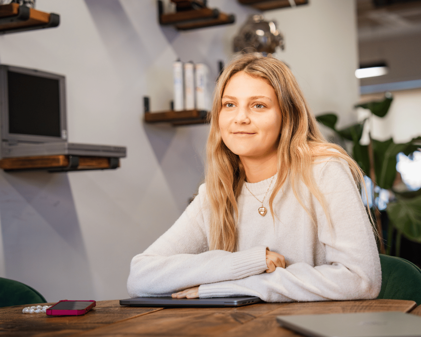 A young woman with long, blonde hair sits at a wooden table in an office, smiling. Plants are in the background.