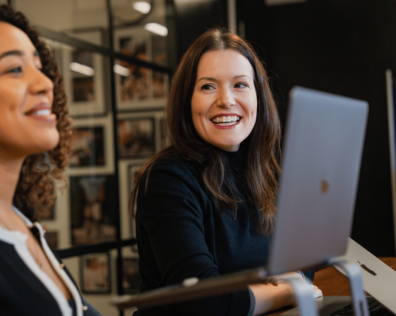 Two women smiling while seated at a table with a laptop. A collaborative atmosphere in a modern office setting.
