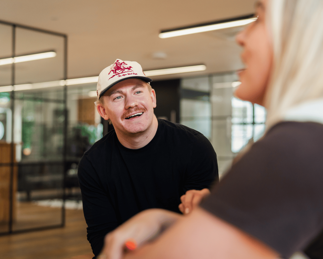 A man in a white cap with a mustache smiles while engaging in conversation with a blurred woman in an office setting