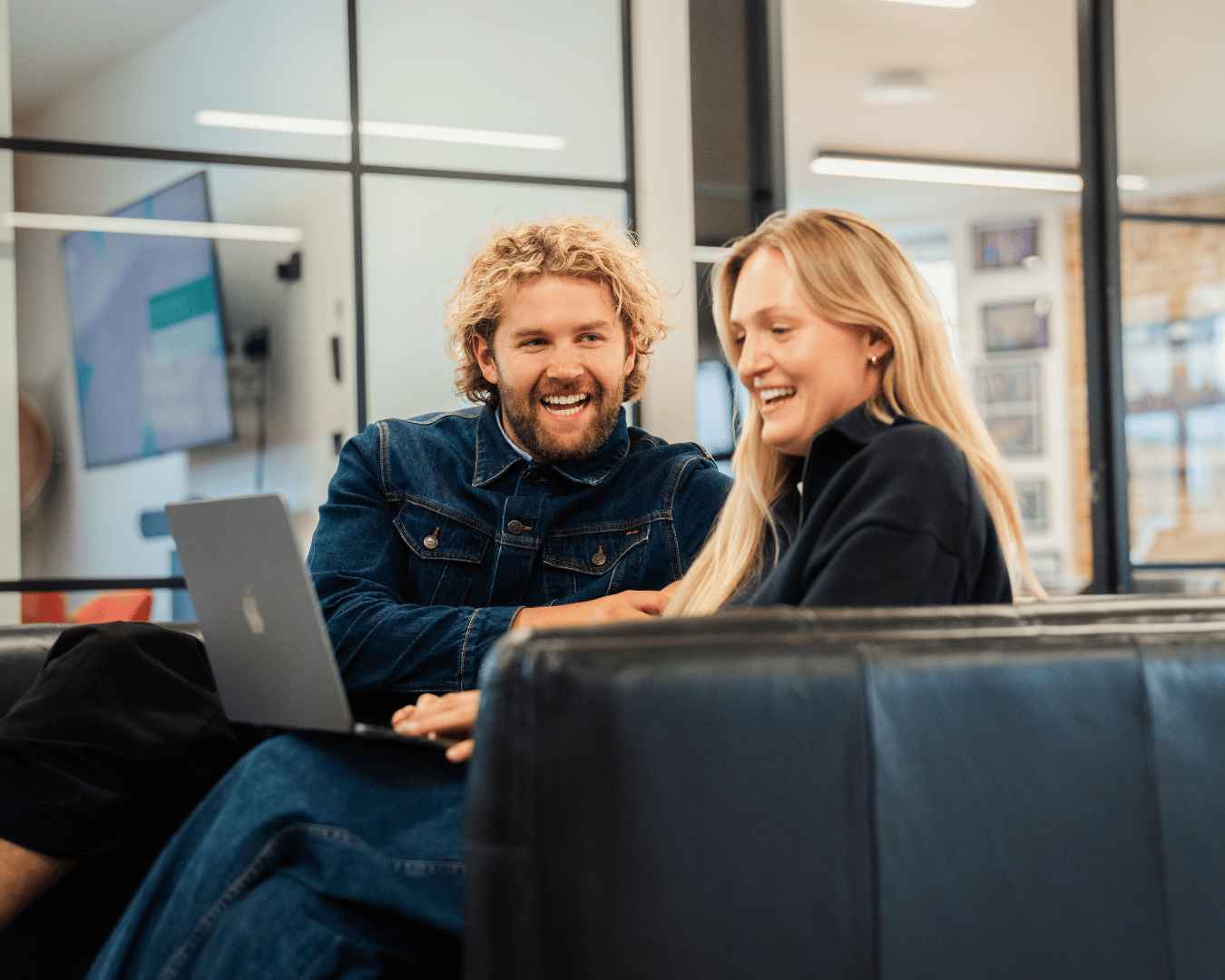 Photo of blonde male and female sat on a office sofa looking at a laptop