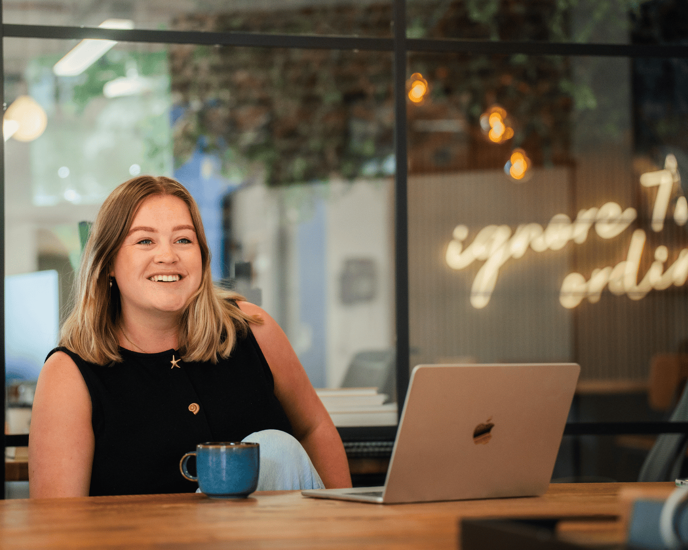 Female employee smiling while working from a laptop in a contemporary office, with a coffee mug on the desk during a team meeting.