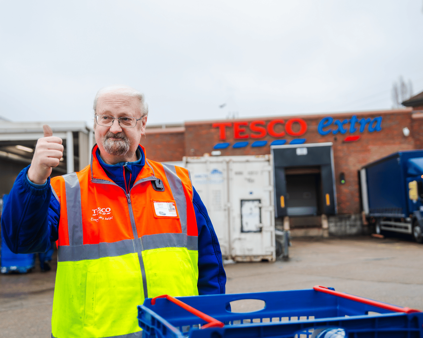 Photo of a man in a supermarket car park wearing an orange and yellow hi-vis jacket with his thumb up