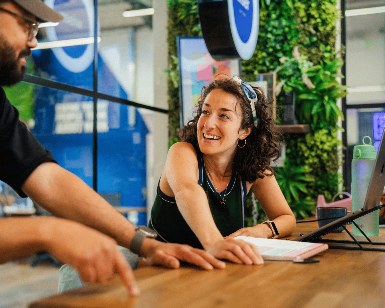 Photo of a female with brown curly hair sat at an office reception desk with greenery in the background