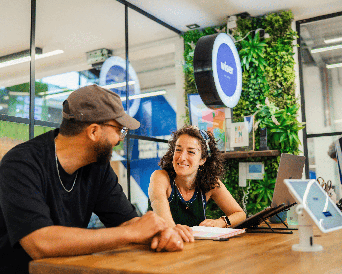 Two smiling coworkers collaborate at a wooden desk in a modern office, with a laptop, tablet, greenery, and a circular wall sign behind them.