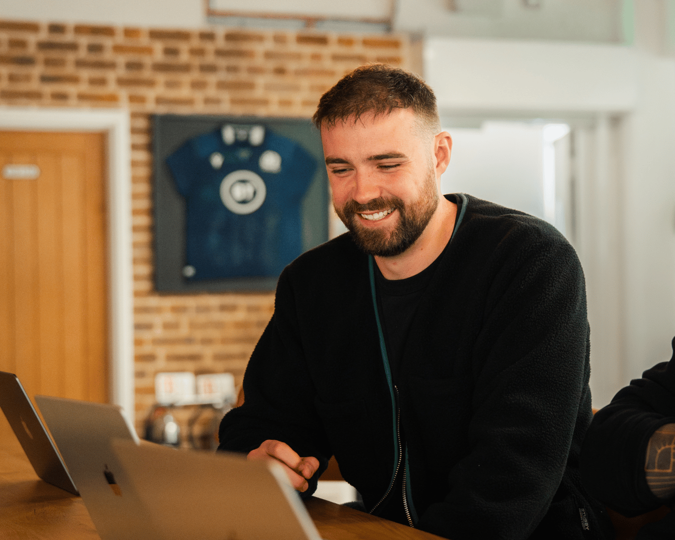 Photo of man with a black jumper smiling looking down at a laptop smiling