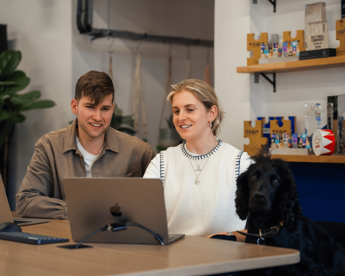 Photo of two people sat at a desk with a dog next to them looking at a laptop and smiling