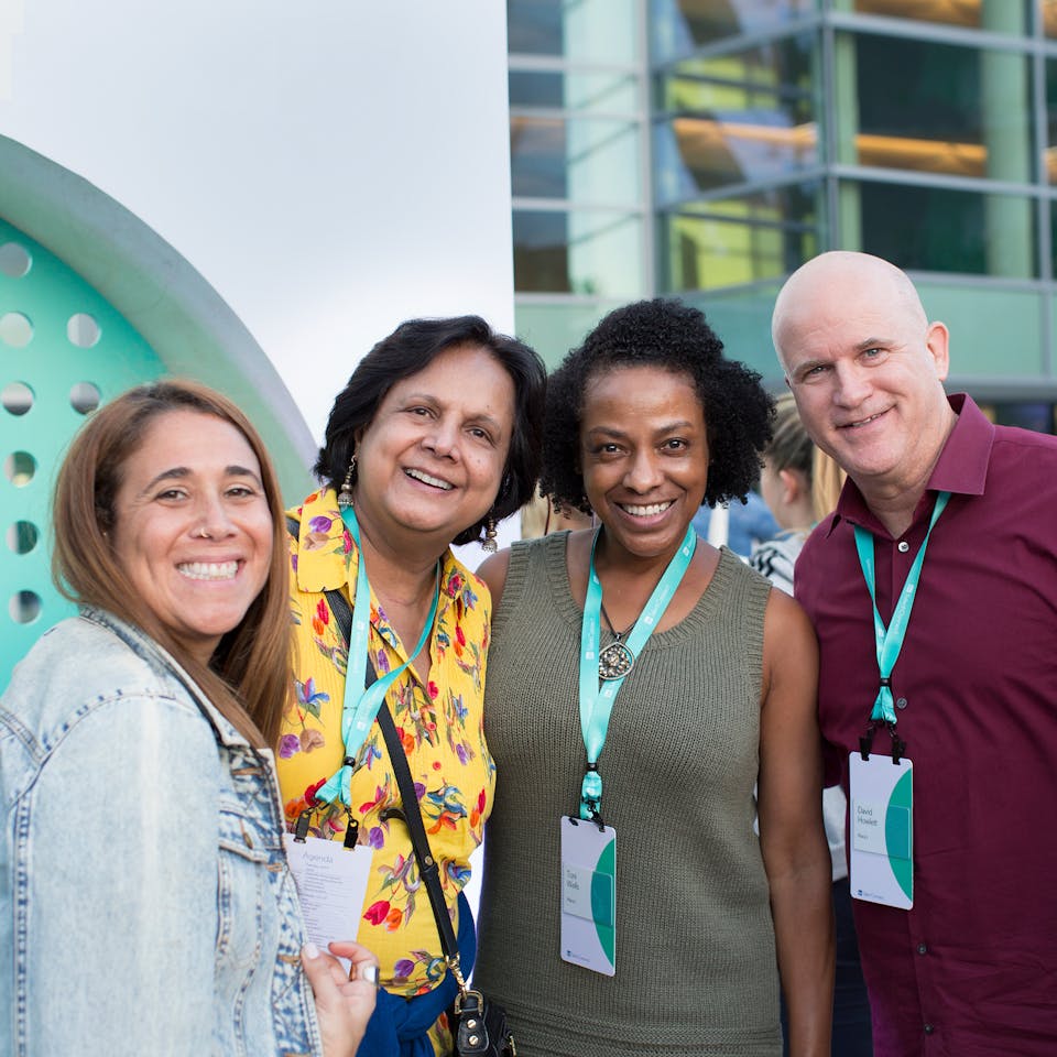Three women and one man smiling with their arms around each other looking forward at the camera.