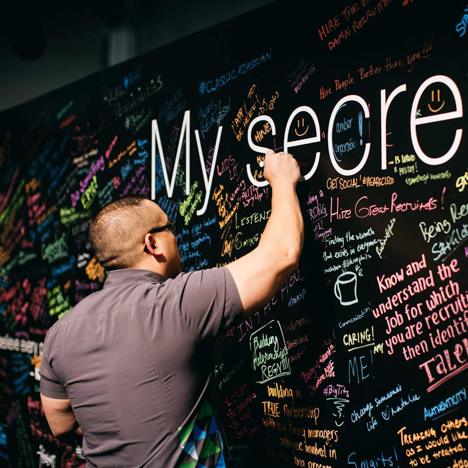 A man in a brown short-sleeved shirt writing on a board that has brightly colored phrases and words.