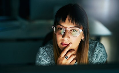 A woman with glasses ponders while looking at a computer screen