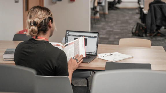 A man sits at a desk with reading a book and an open laptop computer