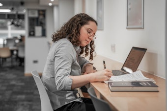 A woman sits at a desk with a computer and writing pad