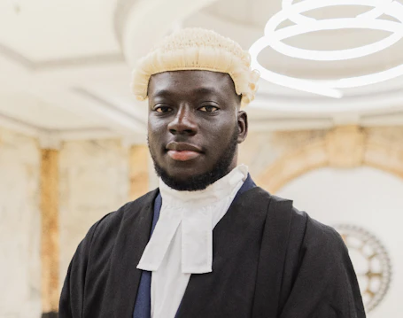 A male barrister stands infront of a stone decorated wall in full gown and wig