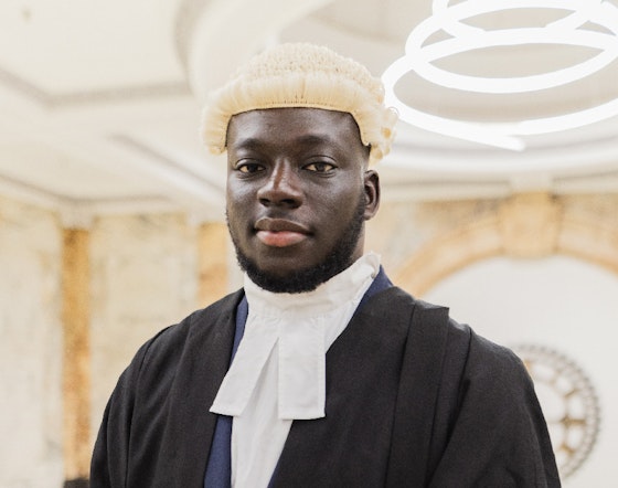 A male barrister stands infront of a stone decorated wall in full gown and wig