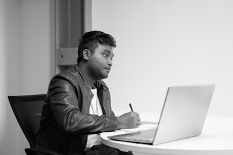 A student writes on a notepad sitting at a desk, a laptop computer is infront of him