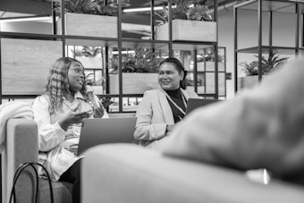 Two female students sit on a sofa with laptops