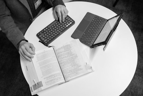 A tablet computer and textbook sits on a white desk with a student typing on an external keyboard