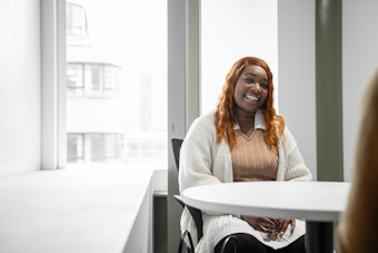 A young woman smiles as she sits across a white desk from a lecturer