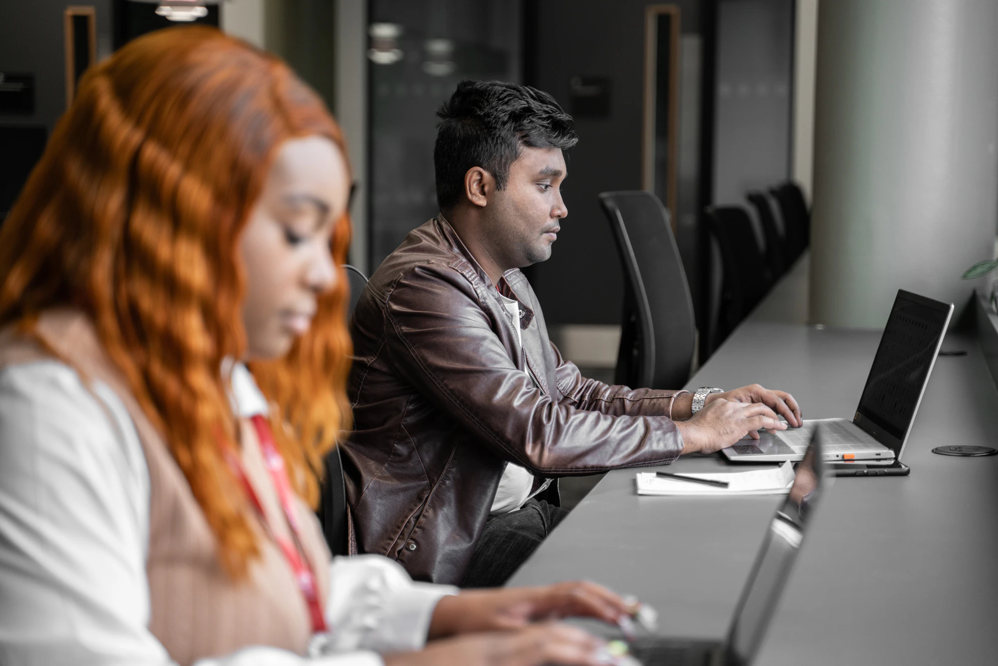A male and female learner sit at a grey desk looking at laptop computers