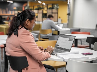 A woman in a pink jacket sits at a laptop in a a brightly-lit BPP student area