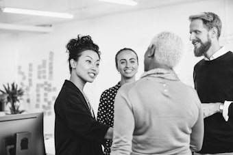 Three females and a male have a conversation while standing in an office