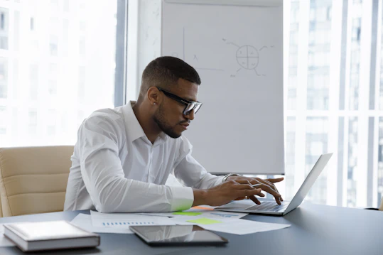 A man wearing glasses sits at a desk typing on a laptop computer