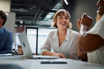 A woman smiles as she converses with a colleague while sitting at a desk with an open laptop computer