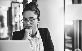 A woman wearing glasses ponders while looking at a laptop screen