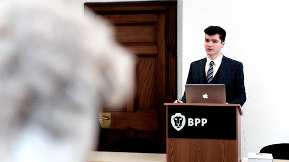 A male stands at lecturn infront of a person wearing a barrister wig