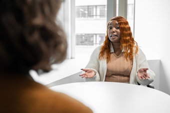 A female student with red hair speaks to a lecturer across a white table