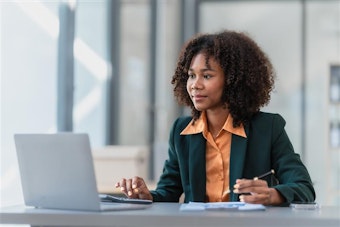 A smartly dressed woman sits at a desk with a pen, calculator and laptop computer
