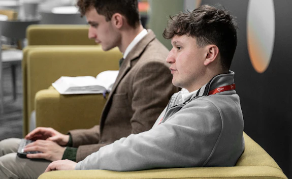 Two male students sit on a green sofa with a laptop and textbook