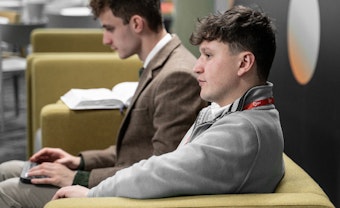 Two male students sit on a green sofa with a laptop and textbook