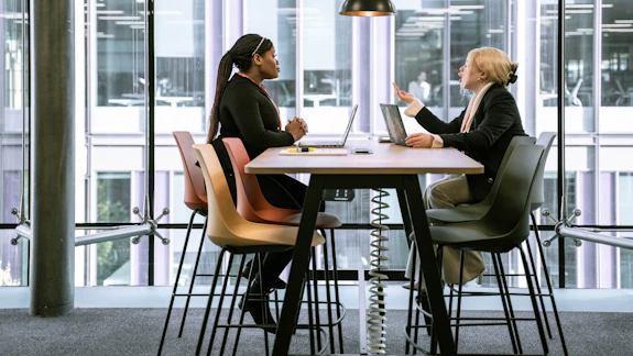 Two students sit across a desk infront of large windows during the day
