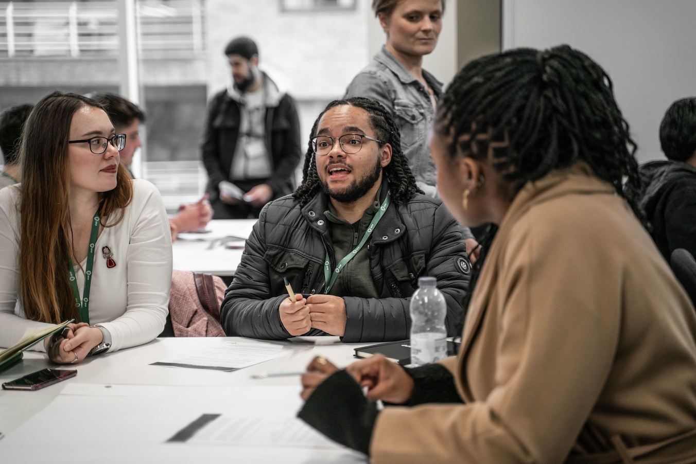 Two students and a member of staff sit a large grey desk containing papers and mobile phones, they are having a conversation.