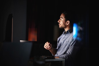 A man in a blue shirt looks at a computer screen in a dark room
