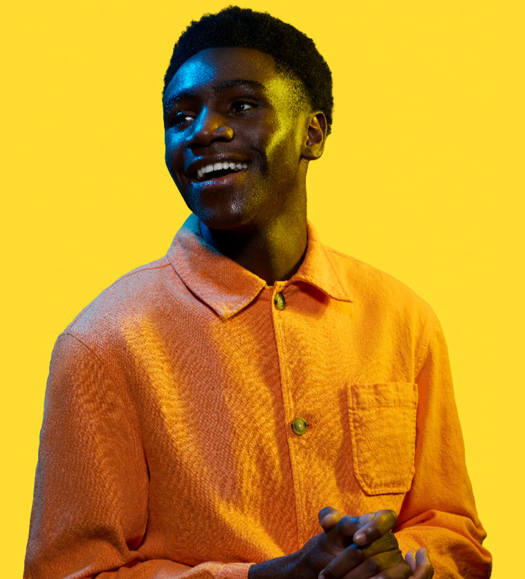 A young man wearing an orange shirt sits with his hands clapsed smiling on a yellow background