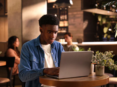 A young man in a white t shirt with blue shirt sits in a coffee shop at a wooden table, he is working attentively on a silver-coloured laptop