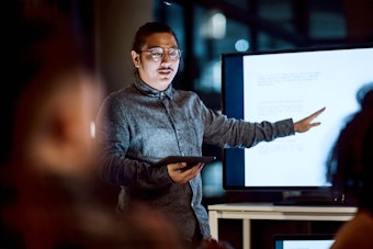 A presenter standing at the front of a room, gesturing toward content displayed on a large digital screen while holding a tablet.