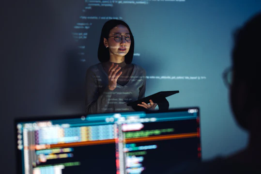 Facilitator with a tablet code Person standing in front of a screen with technical text, holding a tablet while speaking to others in a dimly lit workspace.