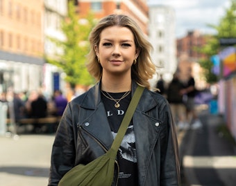Jess Foster stands in a street on a bright day wearing a black leather jacket and green bag