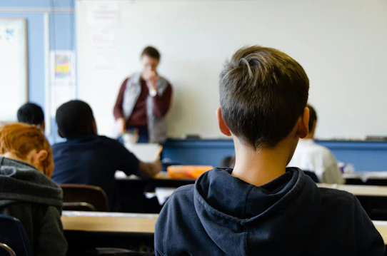 A group of school children sit in class as a teacher presents next to a whiteboard