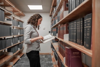 A woman reads an open book while standing in the middle of two aisles of books in a library