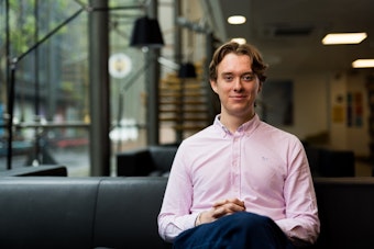 Person sitting on a sofa in a modern office lounge, wearing a light pink shirt and facing the camera, with large windows and a staircase visible in the background.