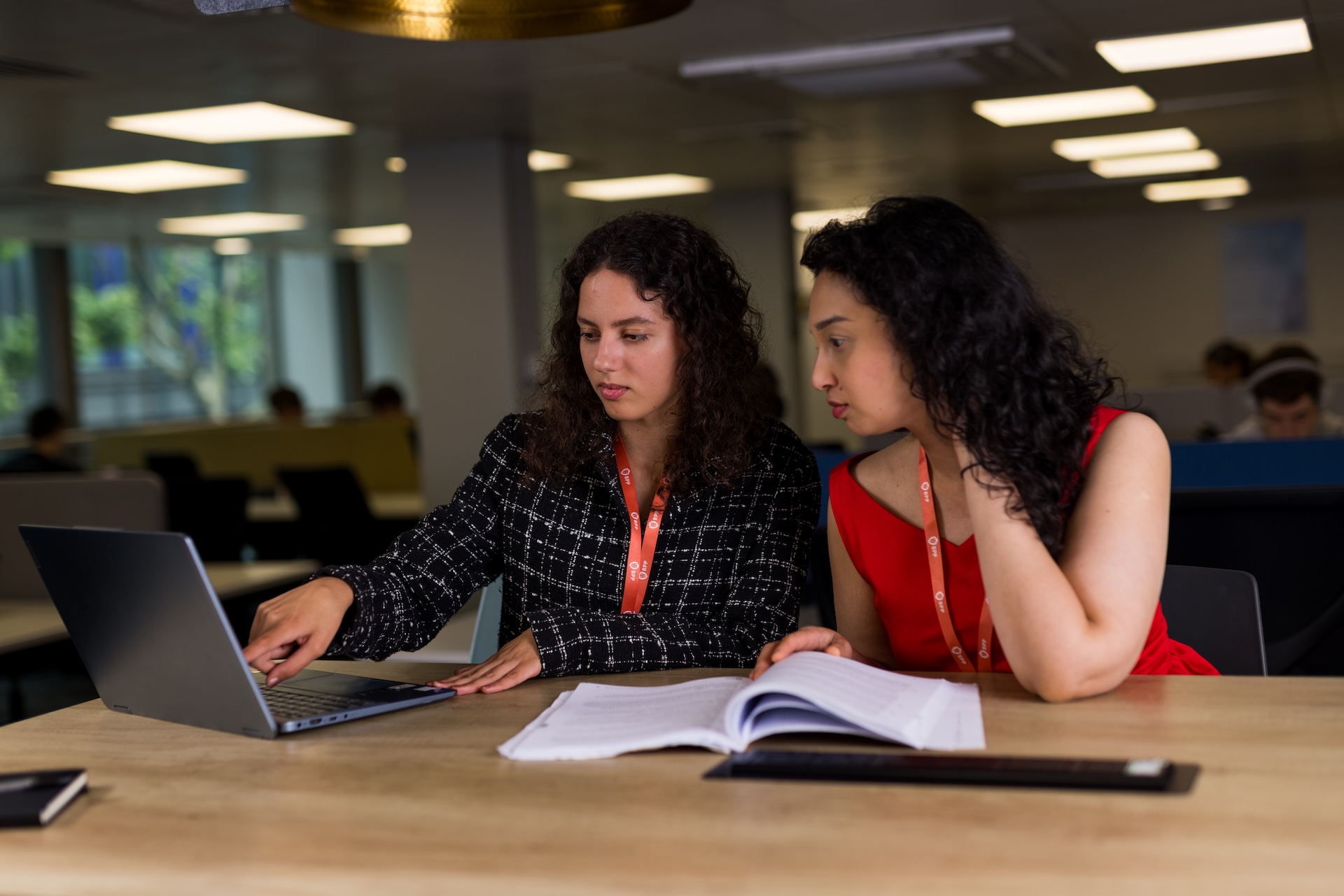 Two learners sit at a table in a study space, reviewing an open book while one points to content on a laptop screen. Both wear lanyards, with blurred desks and overhead lighting visible in the background.
