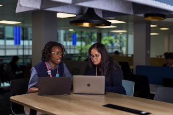 Two people reviewing work on laptops at a shared table in a contemporary office, with large windows and workstations in the background.