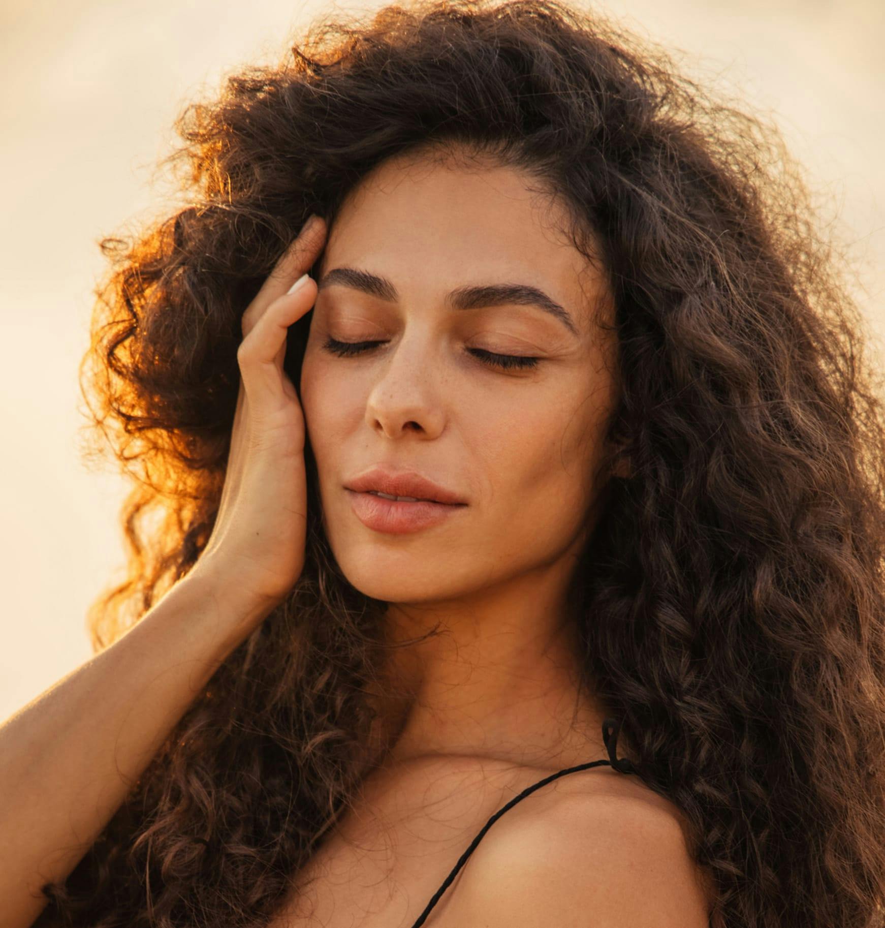 a woman with long curly hair holding her head with sunset in background